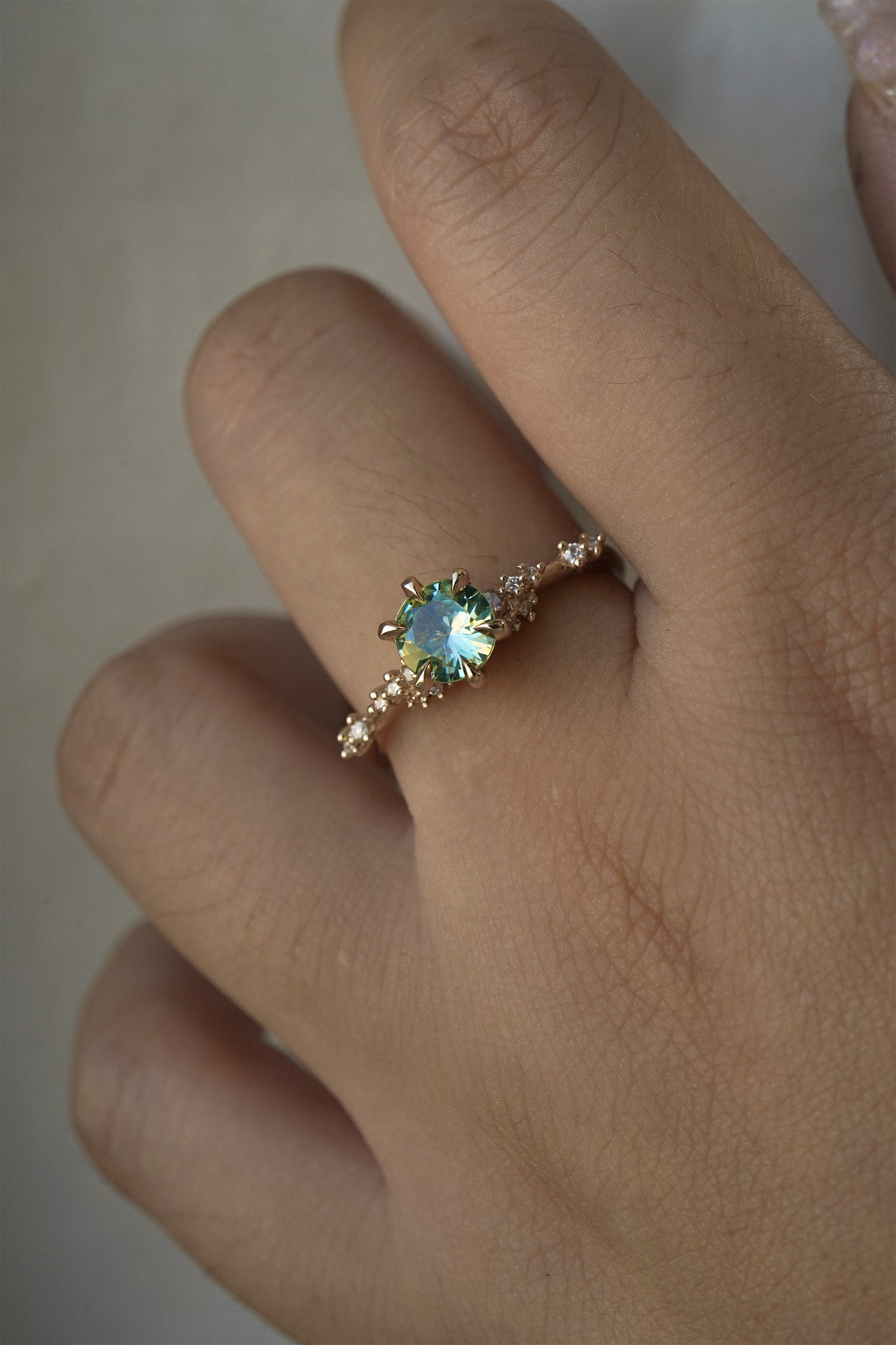 A delicate and ethereal hand-carved one of a kind "Daphne" style engagement ring by Laurie Fleming Jewellery. The ring features a round brilliant cut light green sapphire centre, with clusters of rose and brilliant cut diamond accents on the band in 14k yellow gold. The ring is worn on a hand against a light grey background.
