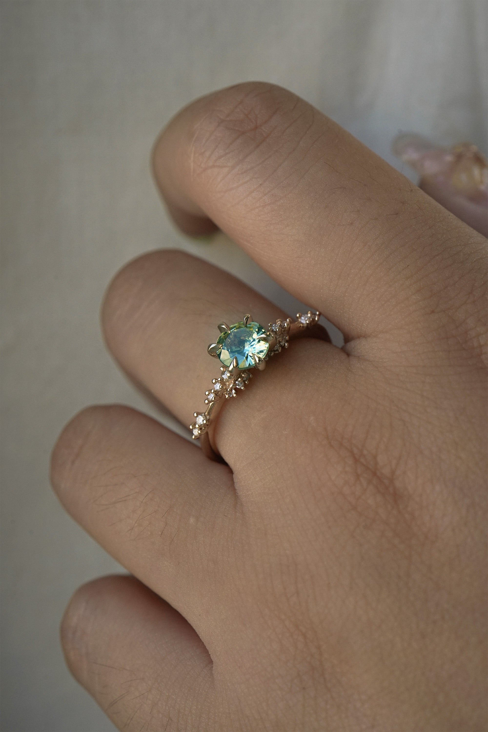 A delicate and ethereal hand-carved one of a kind "Daphne" style engagement ring by Laurie Fleming Jewellery. The ring features a round brilliant cut light green sapphire centre, with clusters of rose and brilliant cut diamond accents on the band in 14k yellow gold. The ring is worn on a hand against a light grey background.
