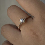 A one of a kind 14k champagne gold "Water Lily" engagement ring by Laurie Fleming Jewellery with a silver-blush colour change Montana sapphire centre held in delicate hand-carved claw prongs. A sprinkling of petite and delicate diamonds dance along the band. The ring is worn on a hand against a light grey background.