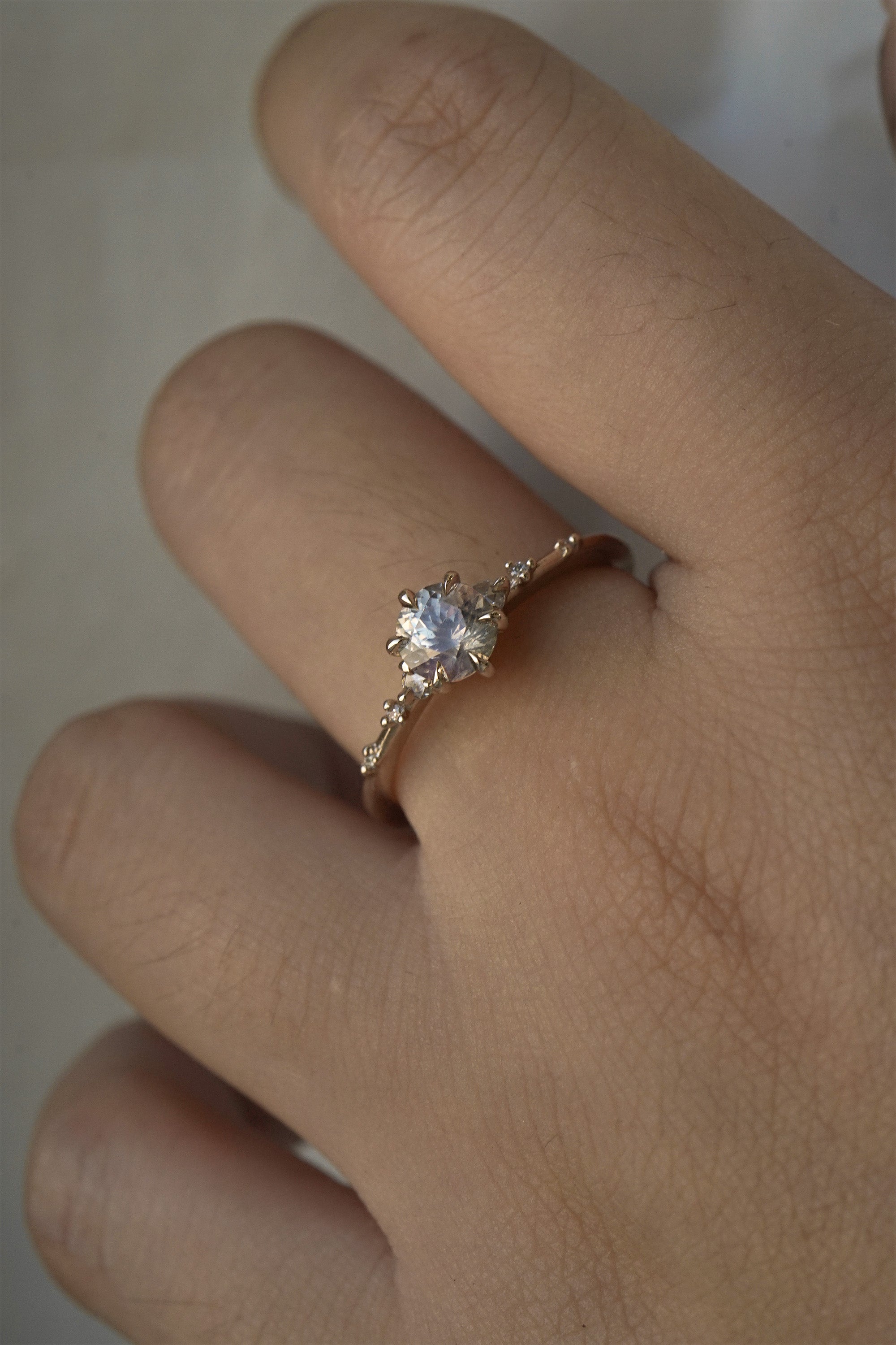 A one of a kind 14k champagne gold "Water Lily" engagement ring by Laurie Fleming Jewellery with a silver-blush colour change Montana sapphire centre held in delicate hand-carved claw prongs. A sprinkling of petite and delicate diamonds dance along the band. The ring is worn on a hand against a light grey background.