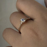 A one of a kind 14k champagne gold "Water Lily" engagement ring by Laurie Fleming Jewellery with a silver-blush colour change Montana sapphire centre held in delicate hand-carved claw prongs. A sprinkling of petite and delicate diamonds dance along the band. The ring is worn on a hand against a light grey background.