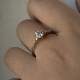A one of a kind 14k champagne gold "Water Lily" engagement ring by Laurie Fleming Jewellery with a silver-blush colour change Montana sapphire centre held in delicate hand-carved claw prongs. A sprinkling of petite and delicate diamonds dance along the band. The ring is worn on a hand against a light grey background.