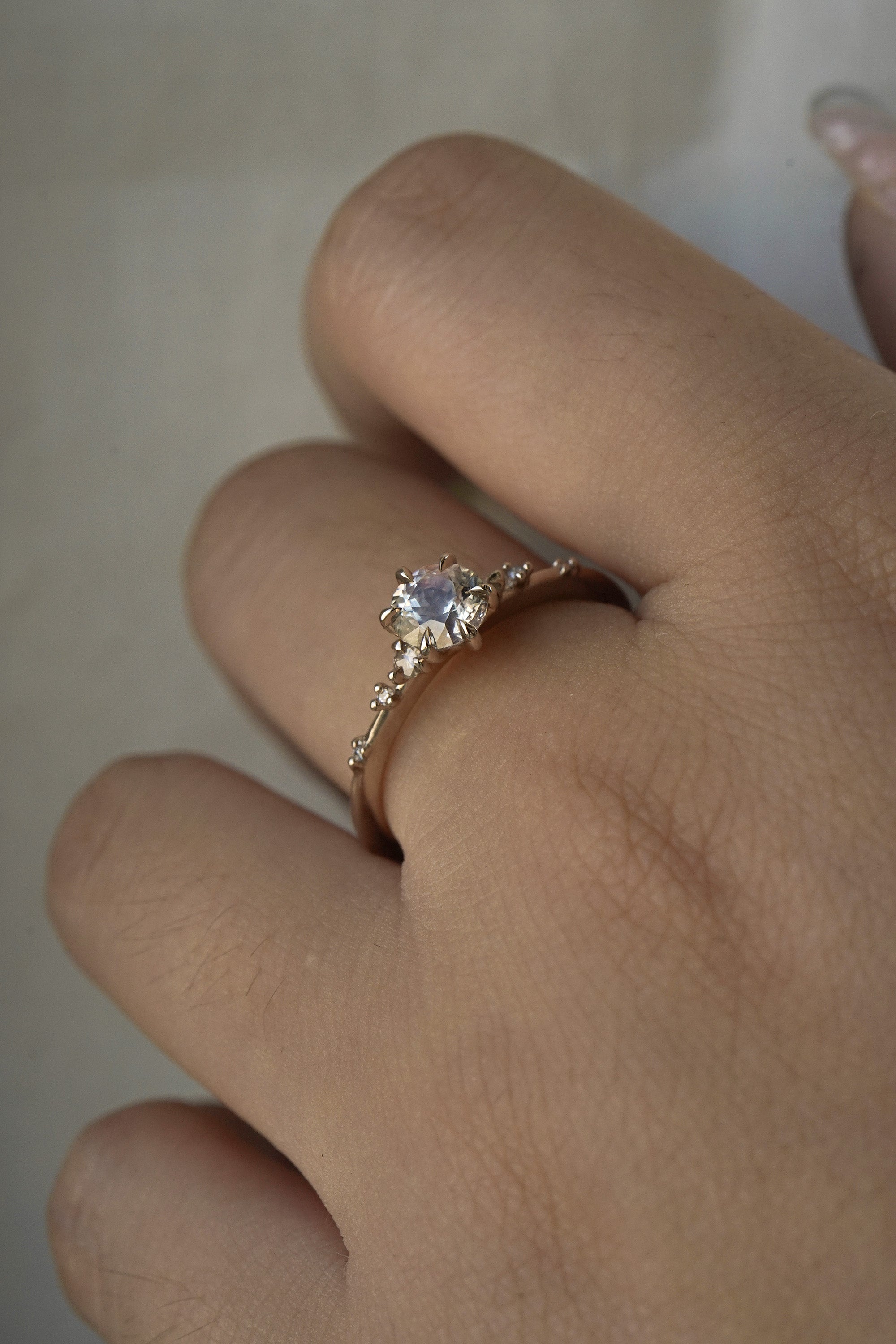 A one of a kind 14k champagne gold "Water Lily" engagement ring by Laurie Fleming Jewellery with a silver-blush colour change Montana sapphire centre held in delicate hand-carved claw prongs. A sprinkling of petite and delicate diamonds dance along the band. The ring is worn on a hand against a light grey background.