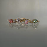 A collection of 4 one of a kind engagement rings by Laurie Fleming Jewellery against a medium grey background. From left to right: a rose gold "Daphne" ring with a vibrant round brilliant cut padparascha sapphire, a "Water Lily" ring with a round brilliant cut silver-blush grey sapphire centre, an "Ilona" ring with a rose cut oval silky pink sapphire, and a "Daphne" ring with a round brilliant cut bright green sapphire centre.