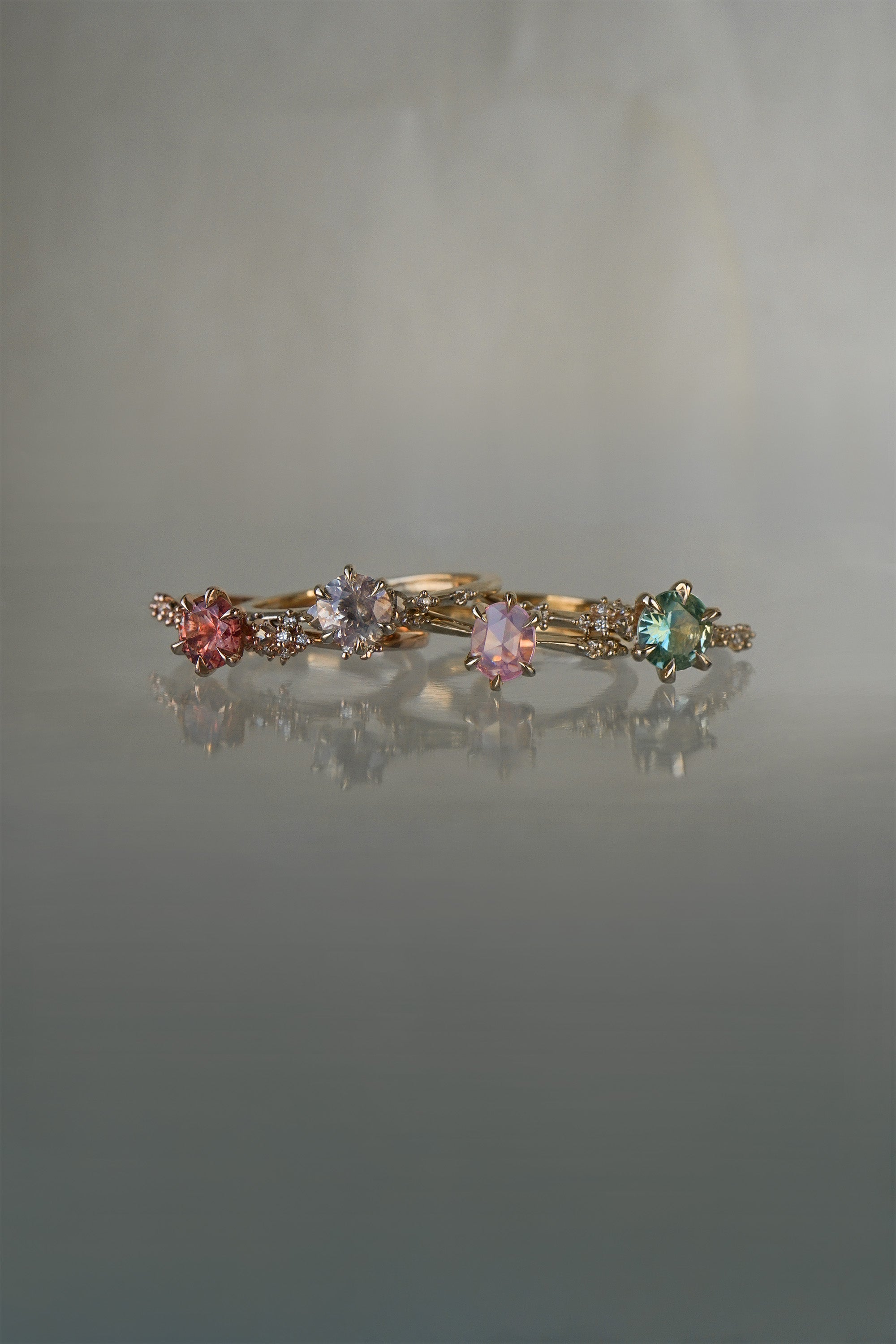 A collection of 4 one of a kind engagement rings by Laurie Fleming Jewellery against a medium grey background. From left to right: a rose gold "Daphne" ring with a vibrant round brilliant cut padparascha sapphire, a "Water Lily" ring with a round brilliant cut silver-blush grey sapphire centre, an "Ilona" ring with a rose cut oval silky pink sapphire, and a "Daphne" ring with a round brilliant cut bright green sapphire centre.