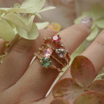 A collection of 4 one of a kind engagement rings by Laurie Fleming Jewellery worn on a hand. From top to bottom: a rose gold "Daphne" ring with a vibrant round brilliant cut padparascha sapphire, a "Water Lily" ring with a round brilliant cut silver-blush grey sapphire centre, an "Ilona" ring with a rose cut oval silky pink sapphire, and a "Daphne" ring with a round brilliant cut bright green sapphire centre. In the foreground, some leaves frame the hand.