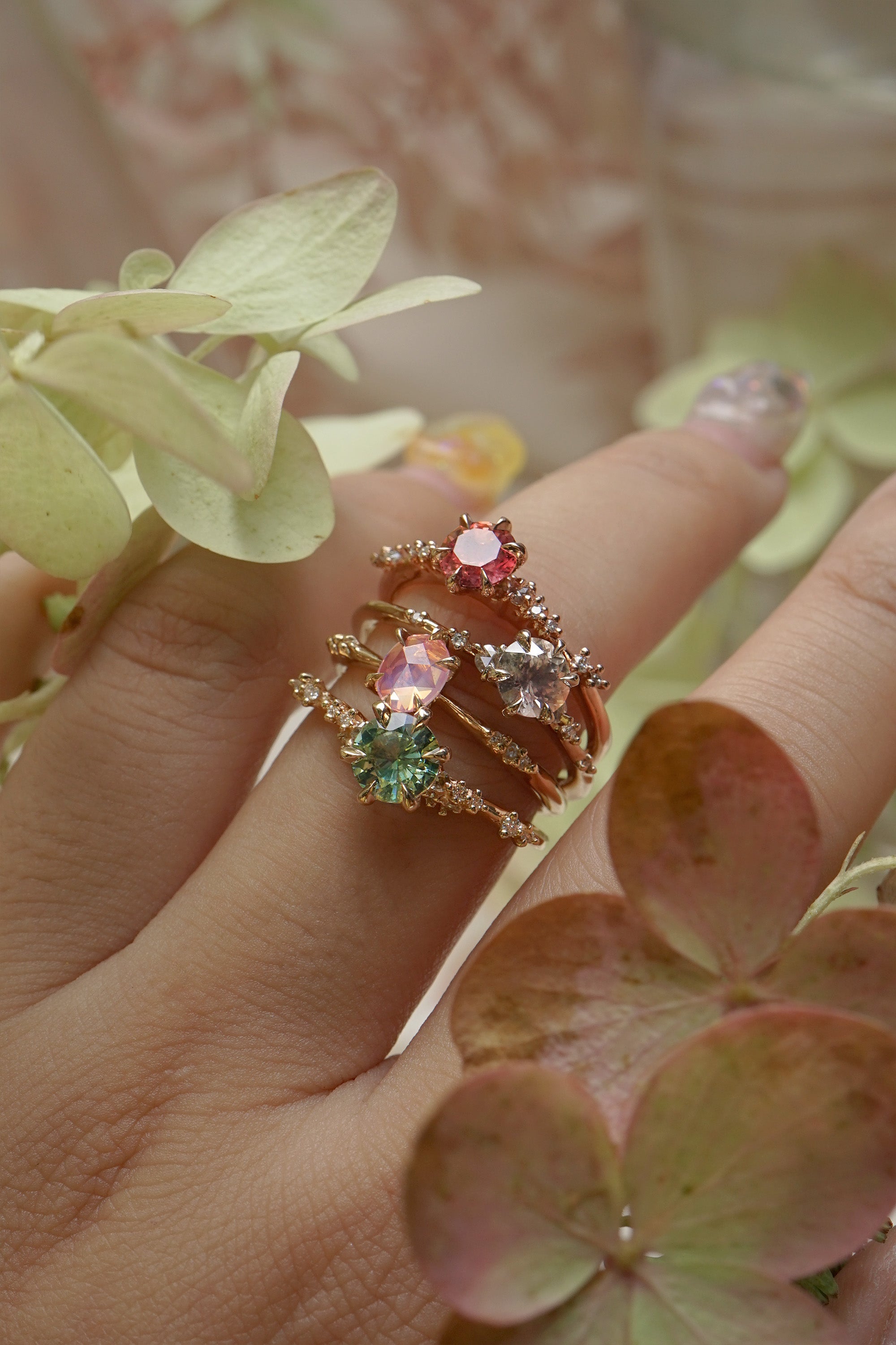 A collection of 4 one of a kind engagement rings by Laurie Fleming Jewellery worn on a hand. From top to bottom: a rose gold "Daphne" ring with a vibrant round brilliant cut padparascha sapphire, a "Water Lily" ring with a round brilliant cut silver-blush grey sapphire centre, an "Ilona" ring with a rose cut oval silky pink sapphire, and a "Daphne" ring with a round brilliant cut bright green sapphire centre. In the foreground, some leaves frame the hand.