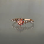 A delicate and ethereal hand-carved one of a kind "Daphne" style engagement ring by Laurie Fleming Jewellery. The ring features a round brilliant cut red peach padparascha sapphire centre, with clusters of rose and brilliant cut diamond accents on the band in 14k rose gold. The ring is situated against a medium grey background.
