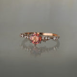A delicate and ethereal hand-carved one of a kind "Daphne" style engagement ring by Laurie Fleming Jewellery. The ring features a round brilliant cut red peach padparascha sapphire centre, with clusters of rose and brilliant cut diamond accents on the band in 14k rose gold. The ring is situated against a medium grey background.