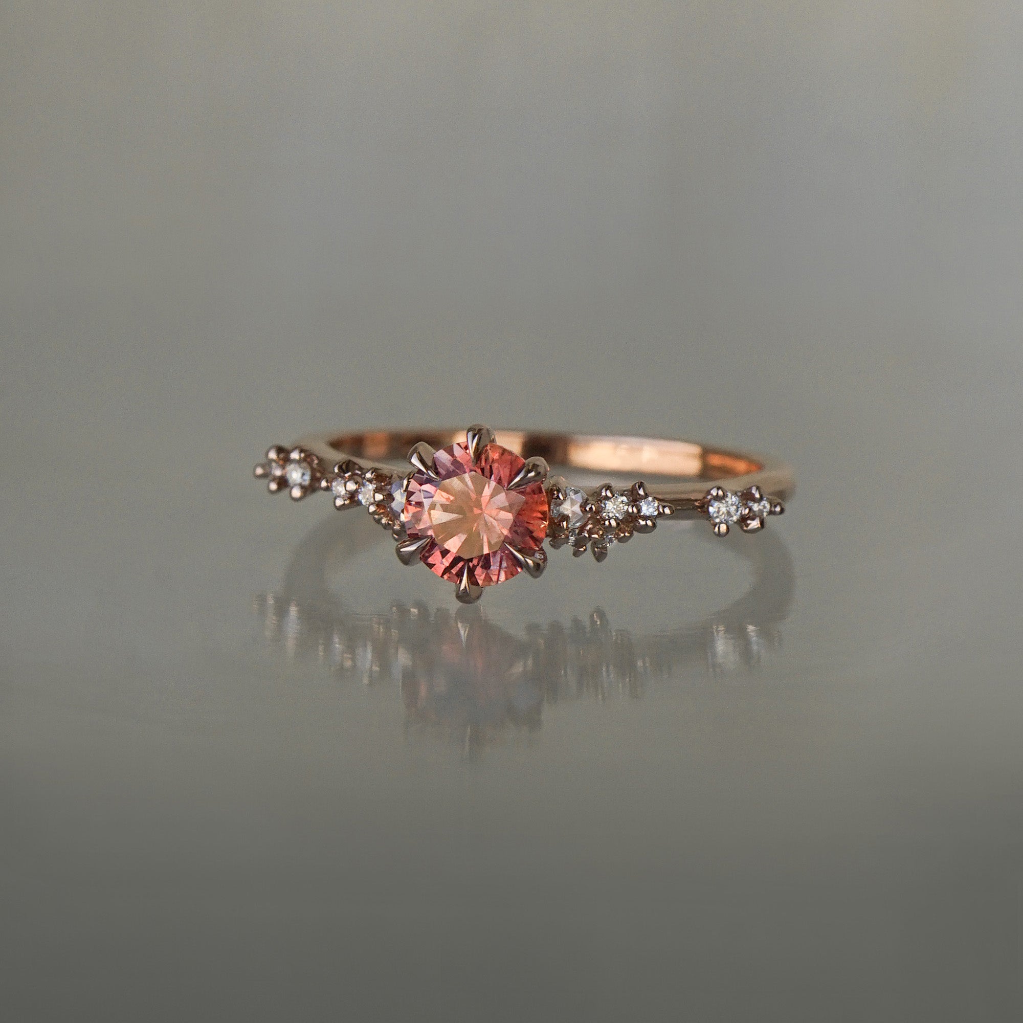 A delicate and ethereal hand-carved one of a kind "Daphne" style engagement ring by Laurie Fleming Jewellery. The ring features a round brilliant cut red peach padparascha sapphire centre, with clusters of rose and brilliant cut diamond accents on the band in 14k rose gold. The ring is situated against a medium grey background.