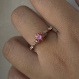A delicate and ethereal hand-carved one of a kind "Daphne" style engagement ring by Laurie Fleming Jewellery. The ring features a round brilliant cut red peach padparascha sapphire centre, with clusters of rose and brilliant cut diamond accents on the band in 14k rose gold. The ring is worn on a hand against a light grey background.