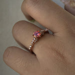 A delicate and ethereal hand-carved one of a kind "Daphne" style engagement ring by Laurie Fleming Jewellery. The ring features a round brilliant cut red peach padparascha sapphire centre, with clusters of rose and brilliant cut diamond accents on the band in 14k rose gold. The ring is worn on a hand against a light grey background.