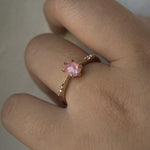 A delicate hand-carved one of a kind "Ilona" style engagement ring by Laurie Fleming Jewellery. The ring features a silky and opalescent rose cut oval pink sapphire centre, a tapered knife-edge band, and small diamond accents on the band shoulders. The ring is worn on a hand against a light grey background.