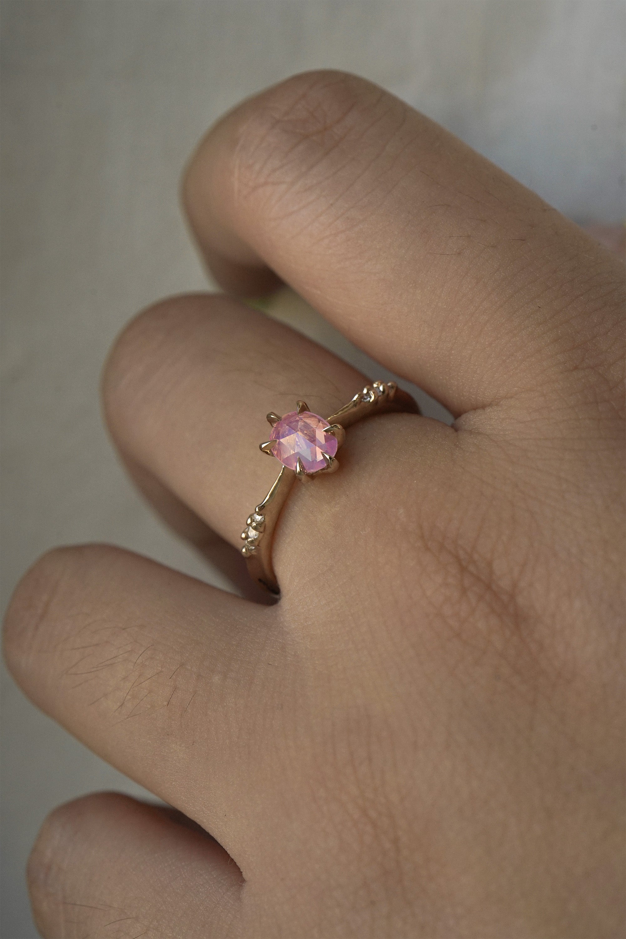 A delicate hand-carved one of a kind "Ilona" style engagement ring by Laurie Fleming Jewellery. The ring features a silky and opalescent rose cut oval pink sapphire centre, a tapered knife-edge band, and small diamond accents on the band shoulders. The ring is worn on a hand against a light grey background.