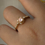 A delicate, one of a kind "Nereid" style engagement ring by Laurie Fleming Jewellery in yellow gold. The ring features a round rose cut light pink sapphire held by four dainty claws, nestled in hand-carved floral peony petals. Rose cut and brilliant diamonds drip down the band. The ring is worn on a hand held in front of a grey background.