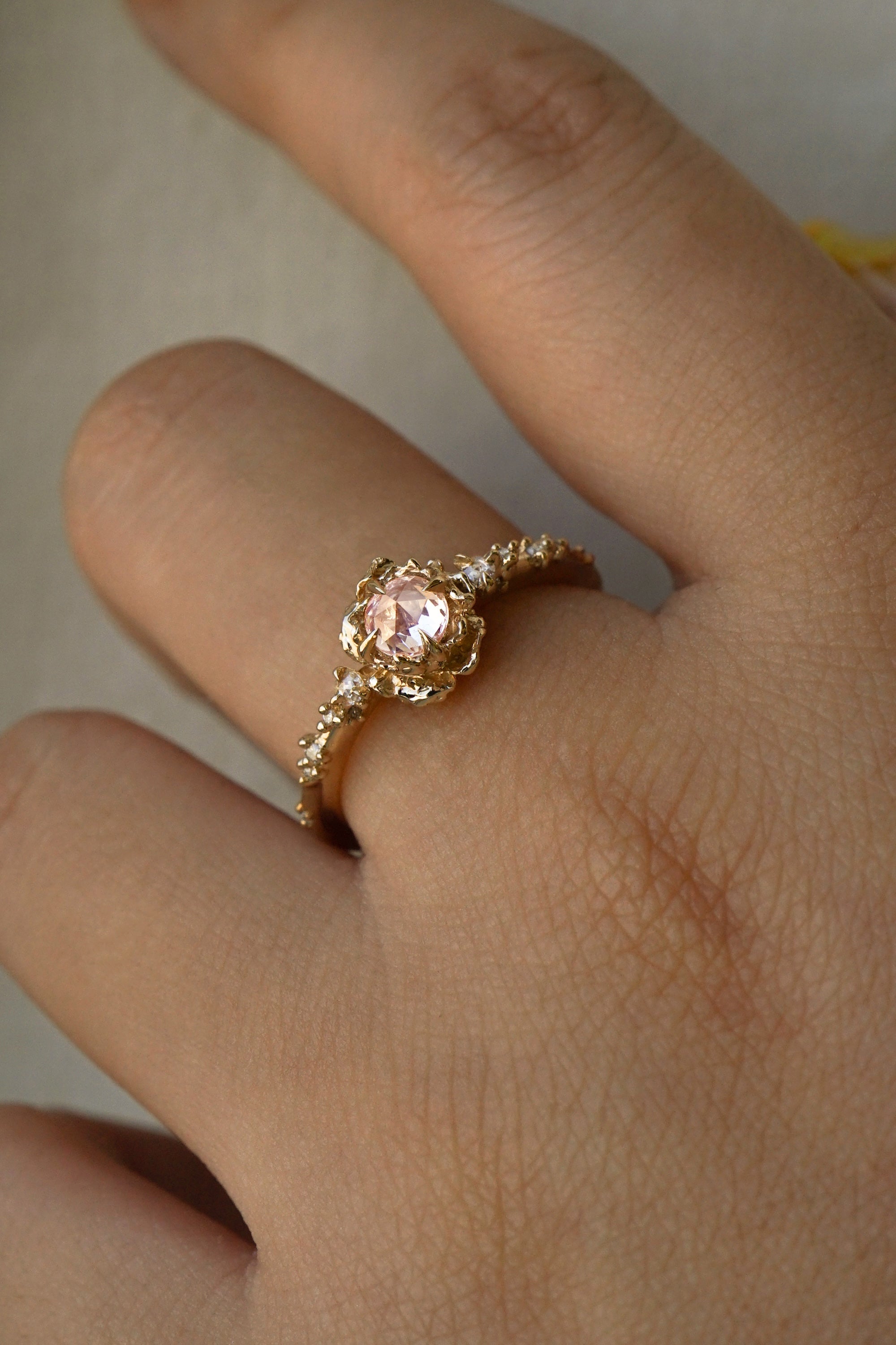 A delicate, one of a kind "Nereid" style engagement ring by Laurie Fleming Jewellery in yellow gold. The ring features a round rose cut light pink sapphire held by four dainty claws, nestled in hand-carved floral peony petals. Rose cut and brilliant diamonds drip down the band. The ring is worn on a hand held in front of a grey background.