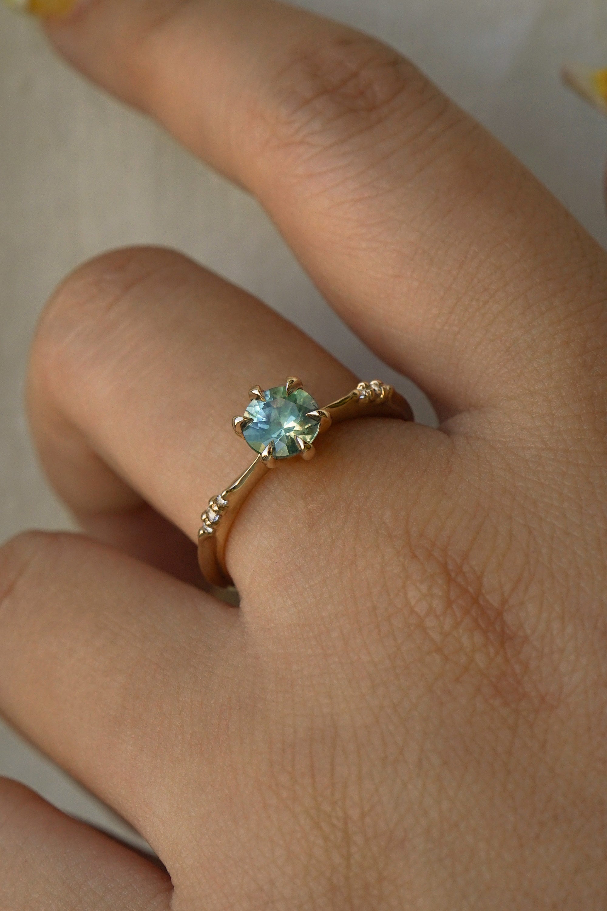 Dainty yellow gold Ilona style one of a kind engagement ring by Laurie Fleming Jewellery, worn on a hand in front of a light grey background. The ring features a silky seafoam turquoise green sapphire and delicate twinkling diamonds on the band.