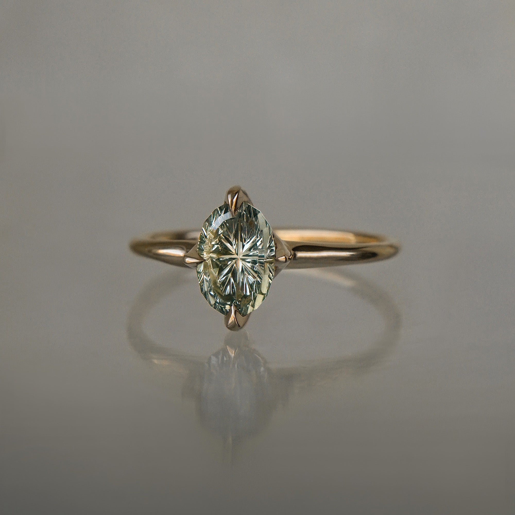 A completely one of a kind, handcarved "Moondew" engagement ring by Laurie Fleming Jewellery. The ring features an oval Starbrite-cut colour change sapphire by lapidary artist John Dyer. The stone is held in four delicate claws, resting atop a gently tapered shining gold band. The ring is situated against a plain grey background.