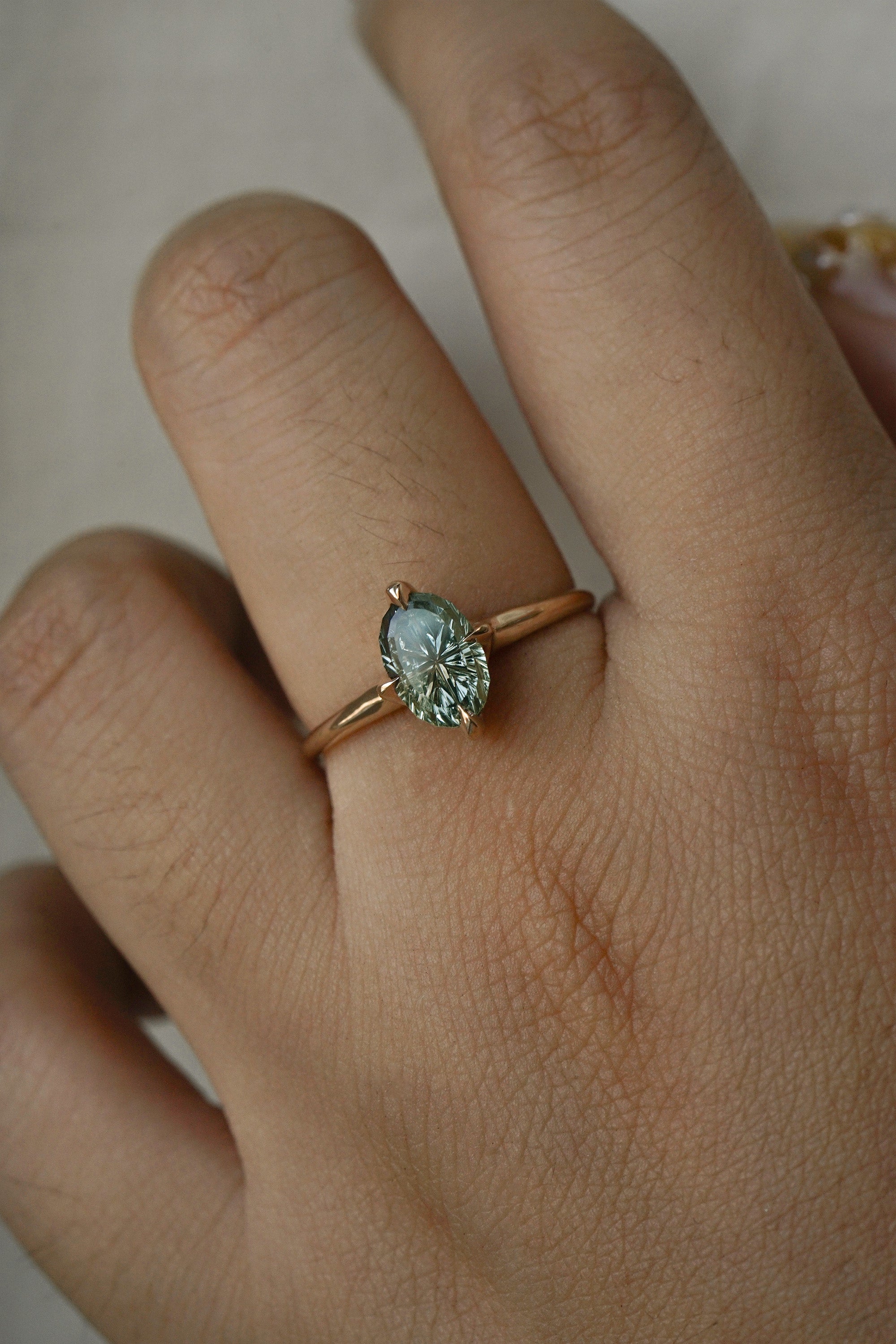 A completely one of a kind, handcarved "Moondew" engagement ring by Laurie Fleming Jewellery. The ring features an oval Starbrite-cut colour change sapphire by lapidary artist John Dyer. The stone is held in four delicate claws, resting atop a gently tapered shining gold band. The ring is worn on a hand against a plain light grey background.