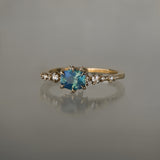A delicate and ethereal "Nereid" one of a kind engagement ring by Laurie Fleming Jewellery, with a unique opalescent silky teal/turquoise/blue sapphire in a geometric oval cut. The band is adorned with rose and brilliant cut diamonds and the ring is made in solid 14k champagne gold. The ring is positioned against a medium grey background.