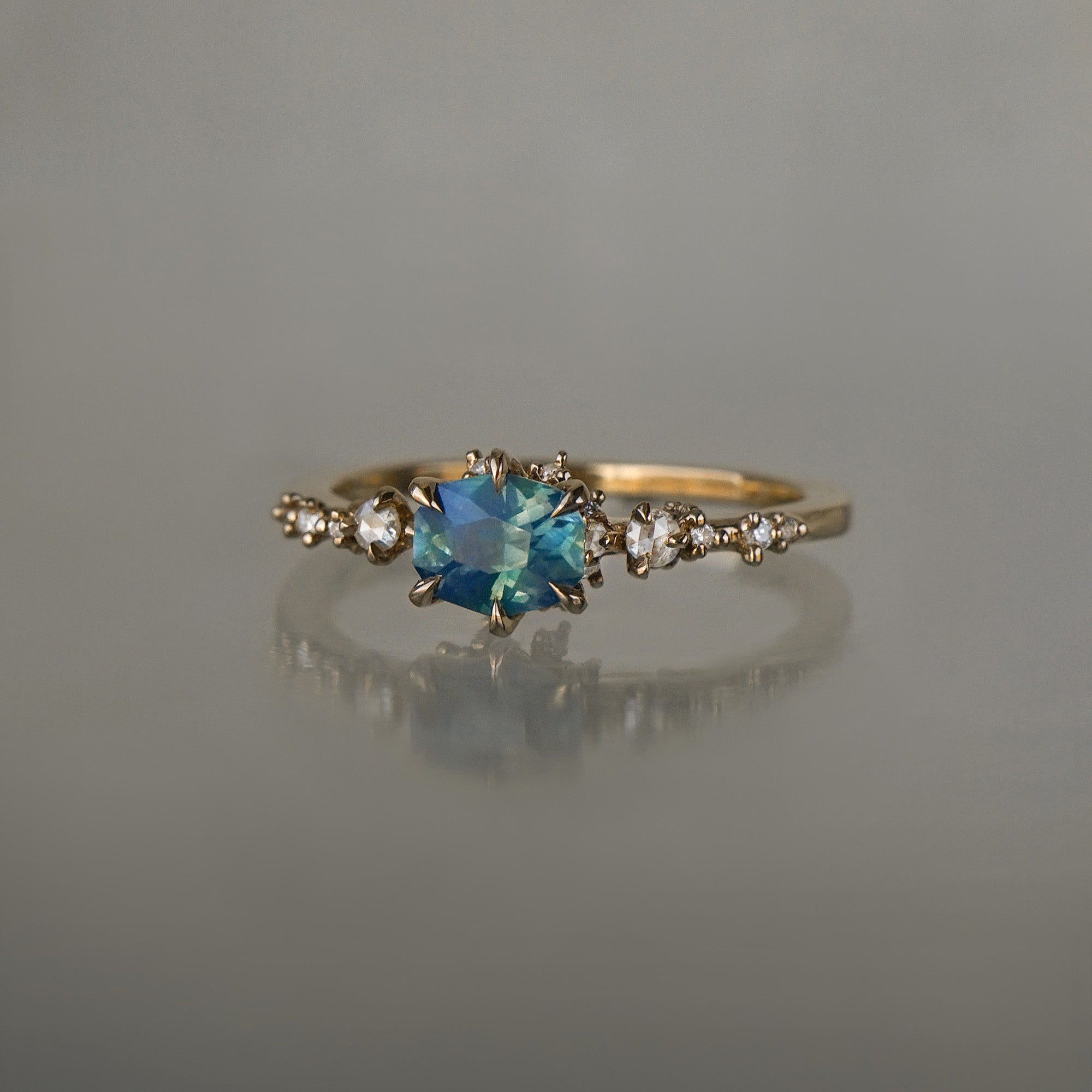 A delicate and ethereal "Nereid" one of a kind engagement ring by Laurie Fleming Jewellery, with a unique opalescent silky teal/turquoise/blue sapphire in a geometric oval cut. The band is adorned with rose and brilliant cut diamonds and the ring is made in solid 14k champagne gold. The ring is positioned against a medium grey background.