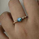 A delicate and ethereal "Nereid" one of a kind engagement ring by Laurie Fleming Jewellery, with a unique opalescent silky teal/turquoise/blue sapphire in a geometric oval cut. The band is adorned with rose and brilliant cut diamonds and the ring is made in solid 14k champagne gold. The ring is worn on a hand against a plain light grey background.