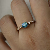 A delicate and ethereal "Nereid" one of a kind engagement ring by Laurie Fleming Jewellery, with a unique opalescent silky teal/turquoise/blue sapphire in a geometric oval cut. The band is adorned with rose and brilliant cut diamonds and the ring is made in solid 14k champagne gold. The ring is worn on a hand against a plain light grey background.