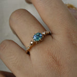 A delicate and ethereal "Nereid" one of a kind engagement ring by Laurie Fleming Jewellery, with a unique opalescent silky teal/turquoise/blue sapphire in a geometric oval cut. The band is adorned with rose and brilliant cut diamonds and the ring is made in solid 14k champagne gold. The ring is worn on a hand against a plain light grey background.