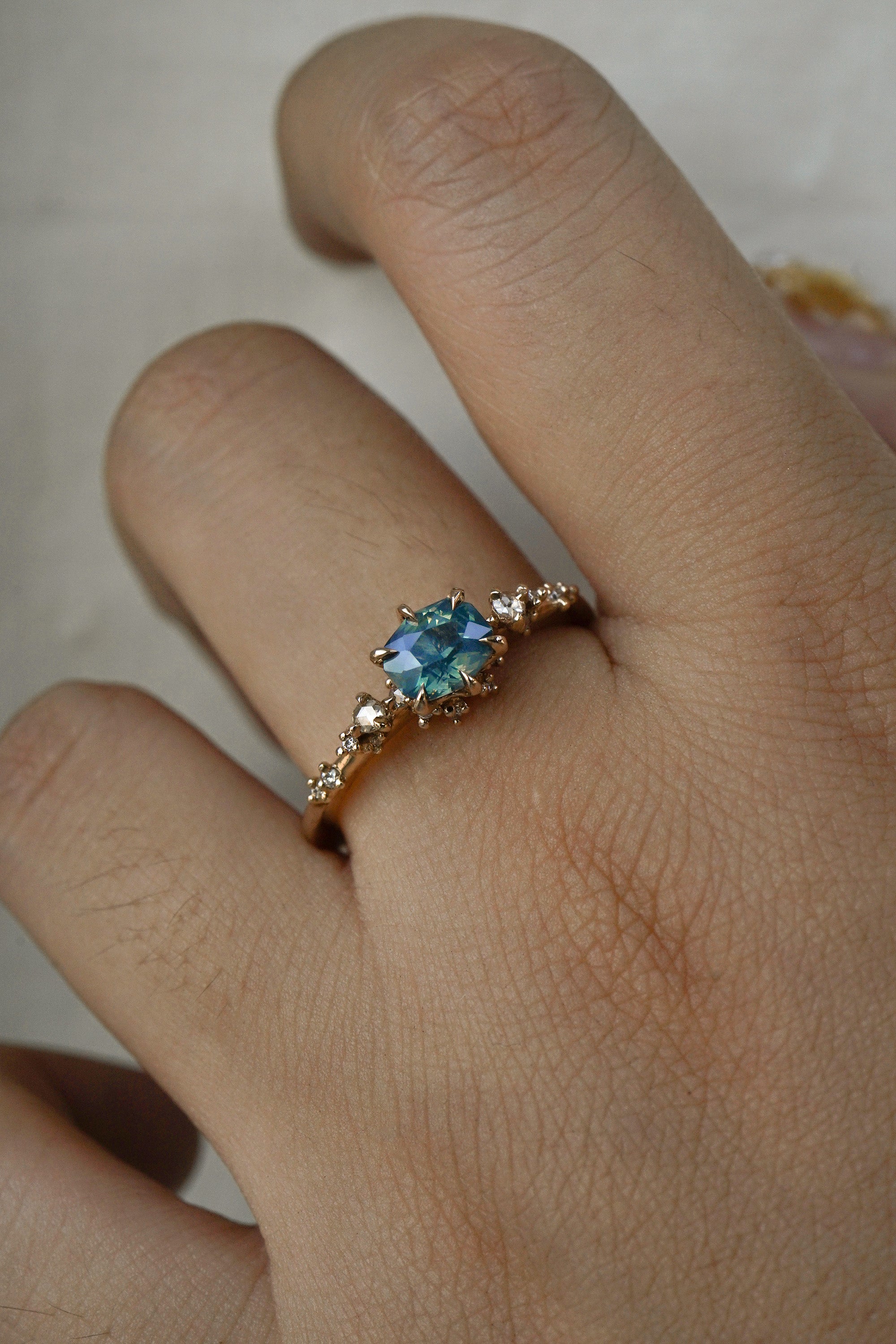 A delicate and ethereal "Nereid" one of a kind engagement ring by Laurie Fleming Jewellery, with a unique opalescent silky teal/turquoise/blue sapphire in a geometric oval cut. The band is adorned with rose and brilliant cut diamonds and the ring is made in solid 14k champagne gold. The ring is worn on a hand against a plain light grey background.