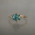 A delicate and ethereal "Nereid" one of a kind engagement ring by Laurie Fleming Jewellery, with a unique opalescent silky teal/turquoise/blue sapphire in an oval cut. The band is adorned with rose and brilliant cut diamonds and the ring is made in solid 14k yellow gold. The ring is positioned against a medium grey background.