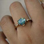 A delicate and ethereal "Nereid" one of a kind engagement ring by Laurie Fleming Jewellery, with a unique opalescent silky teal/turquoise/blue sapphire in an oval cut. The band is adorned with rose and brilliant cut diamonds and the ring is made in solid 14k yellow gold. The ring is worn on a hand against a plain lgiht grey background.