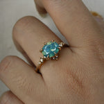 A delicate and ethereal "Nereid" one of a kind engagement ring by Laurie Fleming Jewellery, with a unique opalescent silky teal/turquoise/blue sapphire in an oval cut. The band is adorned with rose and brilliant cut diamonds and the ring is made in solid 14k yellow gold. The ring is worn on a hand against a plain light grey background.