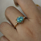 A delicate and ethereal "Nereid" one of a kind engagement ring by Laurie Fleming Jewellery, with a unique opalescent silky teal/turquoise/blue sapphire in an oval cut. The band is adorned with rose and brilliant cut diamonds and the ring is made in solid 14k yellow gold. The ring is worn on a hand against a plain light grey background.