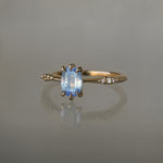 A delicate hand-carved one of a kind "Ilona" style engagement ring by Laurie Fleming Jewellery. The ring features a 0.93ct geometric oval cut silky periwinkle blue sapphire centre, a tapered knife-edge band, and small diamond accents on the band shoulders.  The ring is situated against a medium grey background.