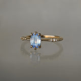 A delicate hand-carved one of a kind "Ilona" style engagement ring by Laurie Fleming Jewellery. The ring features a 0.93ct geometric oval cut silky periwinkle blue sapphire centre, a tapered knife-edge band, and small diamond accents on the band shoulders.  The ring is situated against a medium grey background.