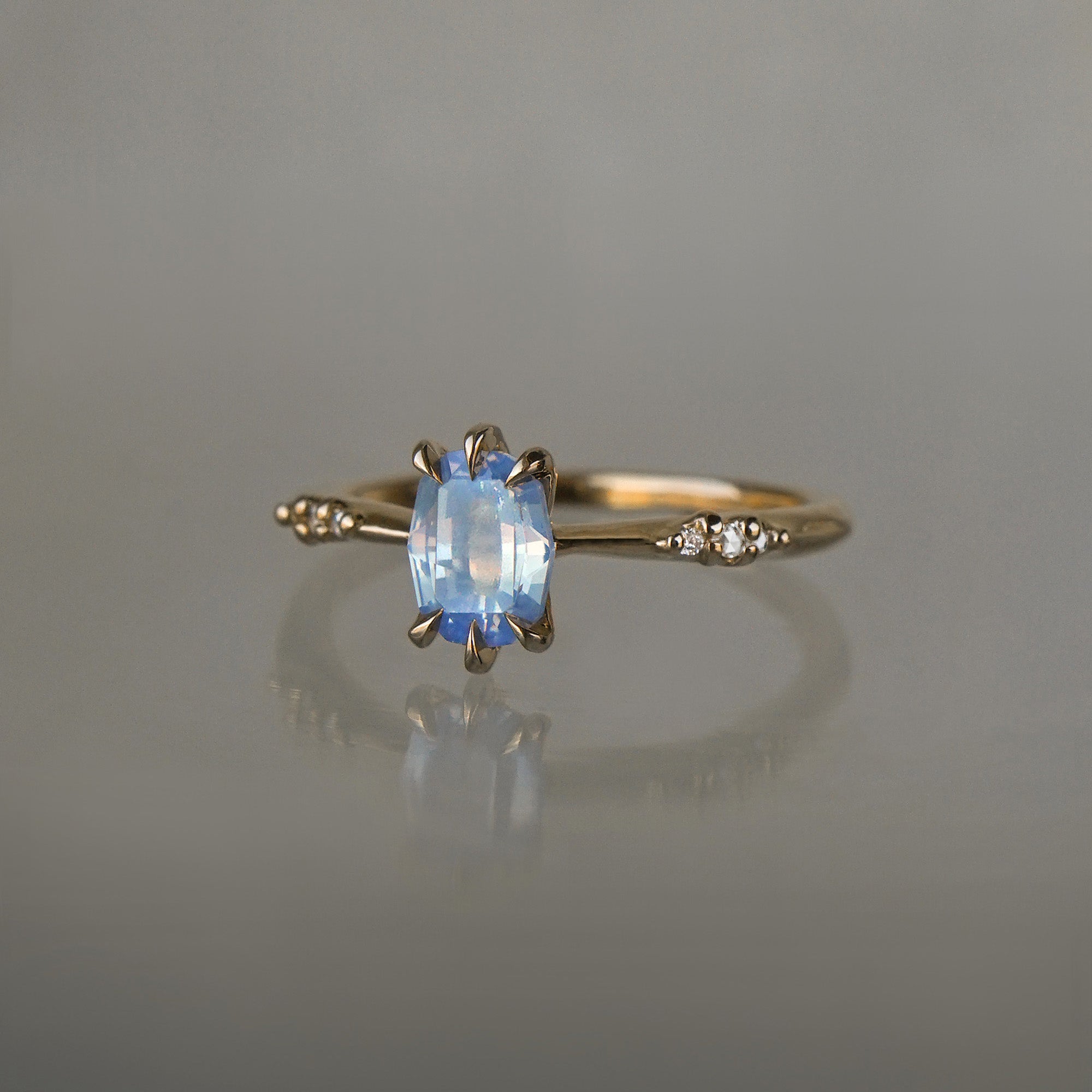 A delicate hand-carved one of a kind "Ilona" style engagement ring by Laurie Fleming Jewellery. The ring features a 0.93ct geometric oval cut silky periwinkle blue sapphire centre, a tapered knife-edge band, and small diamond accents on the band shoulders.  The ring is situated against a medium grey background.