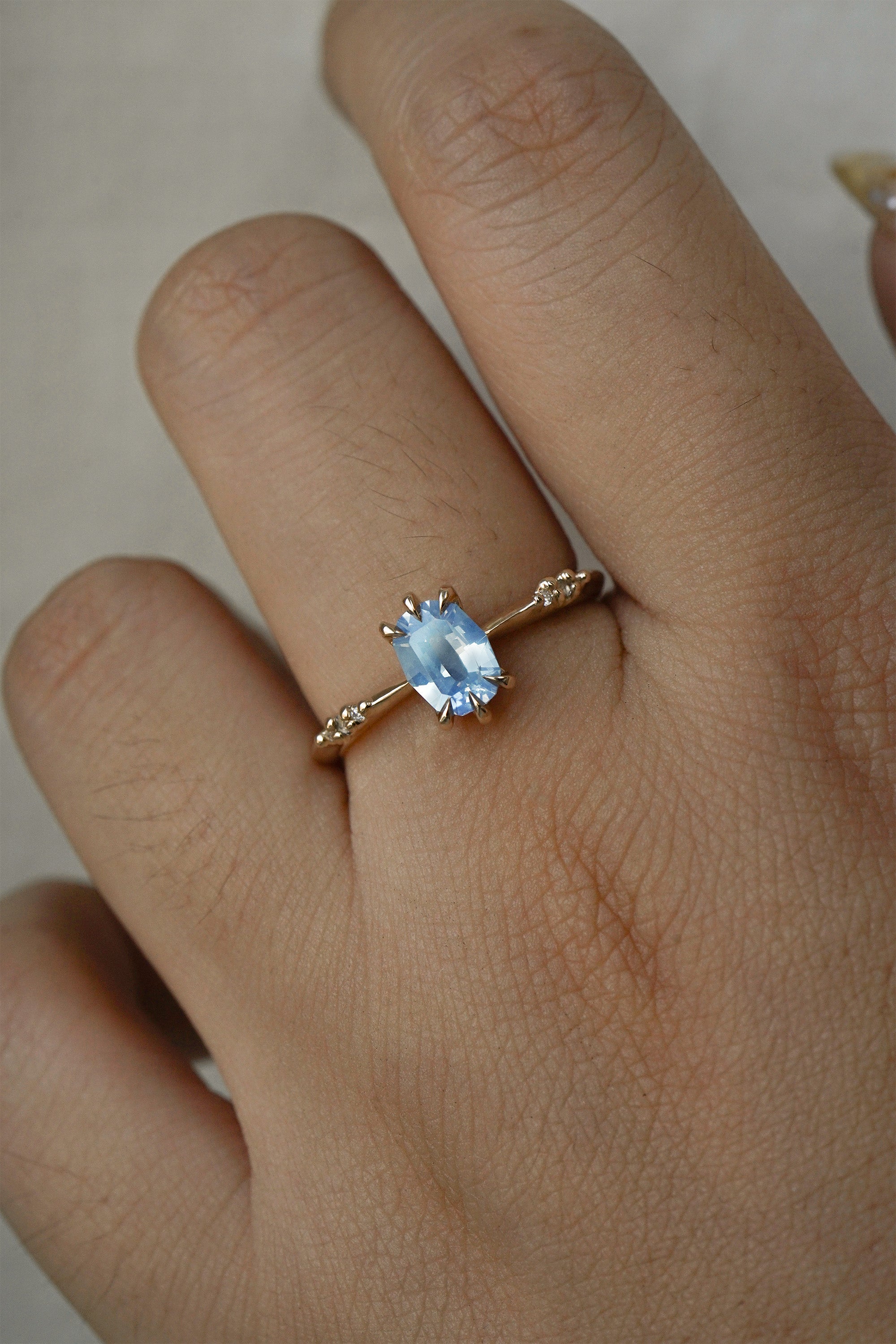 A delicate hand-carved one of a kind "Ilona" style engagement ring by Laurie Fleming Jewellery. The ring features a 0.93ct geometric oval cut silky periwinkle blue sapphire centre, a tapered knife-edge band, and small diamond accents on the band shoulders.  The ring is worn on a hand against a plain light grey background.