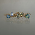 A collection of 5 one of a kind engagement rings by Laurie Fleming Jewellery against a medium grey background. From left to right: a Nereid ring with an opalescent geometric cut blue-green teal sapphire and diamonds on the band, an Ilona ring with a light periwinkle blue oval cut sapphire, a Moondew ring with a colour change Starbrite cut sapphire by John Dyer, an Ilona ring with a yellow-grey round brilliant cut sapphire, and a Nereid ring with a silky opalescent teal oval sapphire.