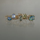 A collection of 5 one of a kind engagement rings by Laurie Fleming Jewellery against a medium grey background. From left to right: a Nereid ring with an opalescent geometric cut blue-green teal sapphire and diamonds on the band, an Ilona ring with a light periwinkle blue oval cut sapphire, a Moondew ring with a colour change Starbrite cut sapphire by John Dyer, an Ilona ring with a yellow-grey round brilliant cut sapphire, and a Nereid ring with a silky opalescent teal oval sapphire.