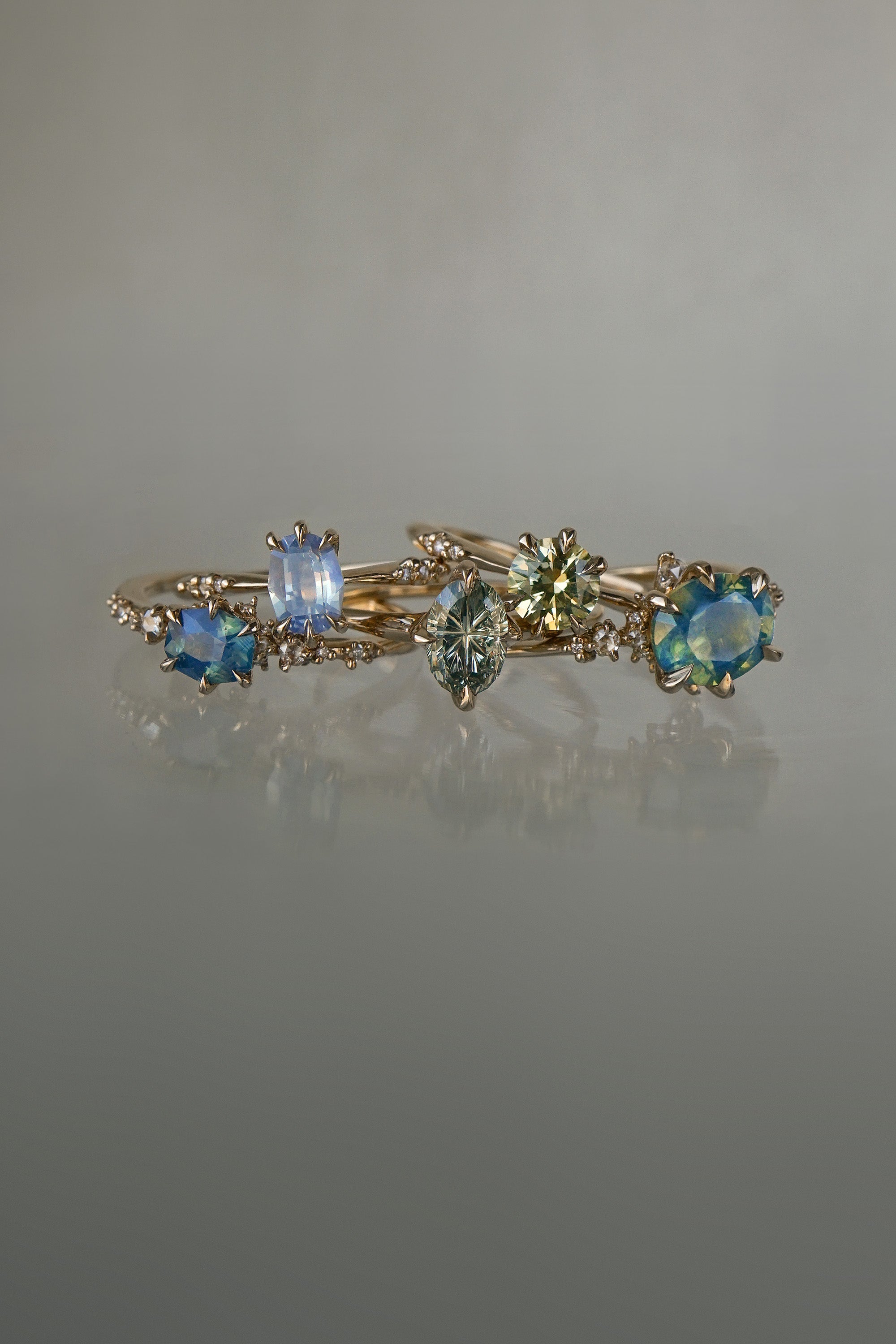 A collection of 5 one of a kind engagement rings by Laurie Fleming Jewellery against a medium grey background. From left to right: a Nereid ring with an opalescent geometric cut blue-green teal sapphire and diamonds on the band, an Ilona ring with a light periwinkle blue oval cut sapphire, a Moondew ring with a colour change Starbrite cut sapphire by John Dyer, an Ilona ring with a yellow-grey round brilliant cut sapphire, and a Nereid ring with a silky opalescent teal oval sapphire.