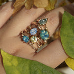 Five one of a kind engagement rings by Laurie Fleming Jewellery worn on a hand, with plants in the fore and background. From left to right: a Nereid ring with an opalescent geometric cut blue-green teal sapphire and diamonds on the band, an Ilona ring with a yellow-grey round brilliant cut sapphire, an Ilona ring with a light periwinkle blue oval cut sapphire, a Nereid ring with a silky opalescent teal oval sapphire, and a Moondew ring with a colour change Starbrite cut sapphire by John Dyer.