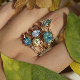 Five one of a kind engagement rings by Laurie Fleming Jewellery worn on a hand, with plants in the fore and background. From left to right: a Nereid ring with an opalescent geometric cut blue-green teal sapphire and diamonds on the band, an Ilona ring with a yellow-grey round brilliant cut sapphire, an Ilona ring with a light periwinkle blue oval cut sapphire, a Nereid ring with a silky opalescent teal oval sapphire, and a Moondew ring with a colour change Starbrite cut sapphire by John Dyer.