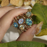 Five one of a kind engagement rings by Laurie Fleming Jewellery held between index and thumb, with plants in the fore and background. From left to right: a Nereid ring with an opalescent geometric cut blue-green teal sapphire and diamonds on the band, an Ilona ring with a yellow-grey round brilliant cut sapphire, an Ilona ring with a light periwinkle blue oval cut sapphire, a Nereid ring with a silky opalescent teal oval sapphire, and a Moondew ring with a colour change Starbrite cut sapphire by John Dyer.
