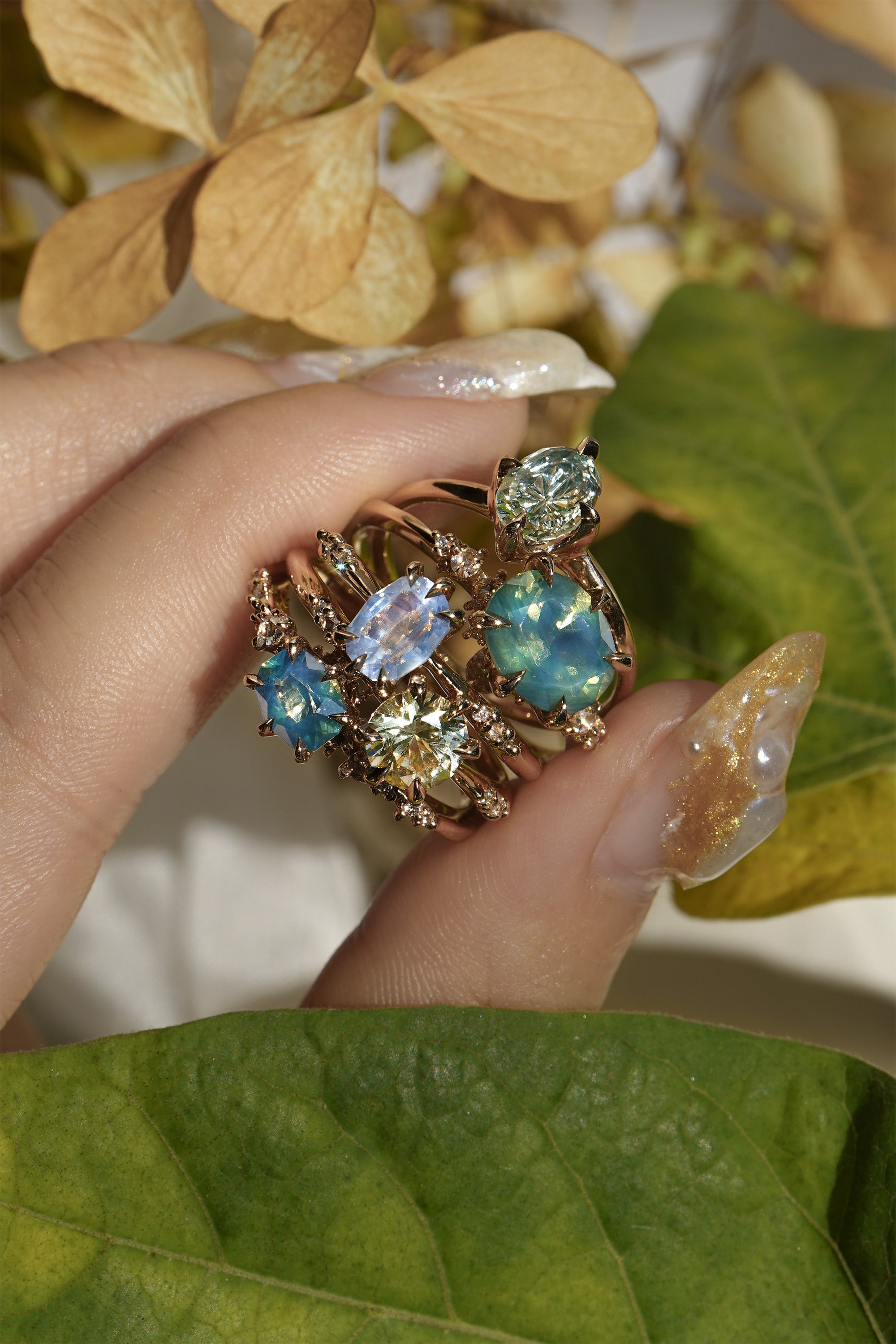 Five one of a kind engagement rings by Laurie Fleming Jewellery held between index and thumb, with plants in the fore and background. From left to right: a Nereid ring with an opalescent geometric cut blue-green teal sapphire and diamonds on the band, an Ilona ring with a yellow-grey round brilliant cut sapphire, an Ilona ring with a light periwinkle blue oval cut sapphire, a Nereid ring with a silky opalescent teal oval sapphire, and a Moondew ring with a colour change Starbrite cut sapphire by John Dyer.