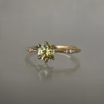 An elegant, one of a kind "Ilona" style engagement ring by Laurie Fleming Jewellery. The ring features a 1ct round brilliant cut yellow-grey Montana sapphire poised atop a tapered knife-edge band with small diamond accents on the band shoulders.  The ring is situated against a medium grey background.