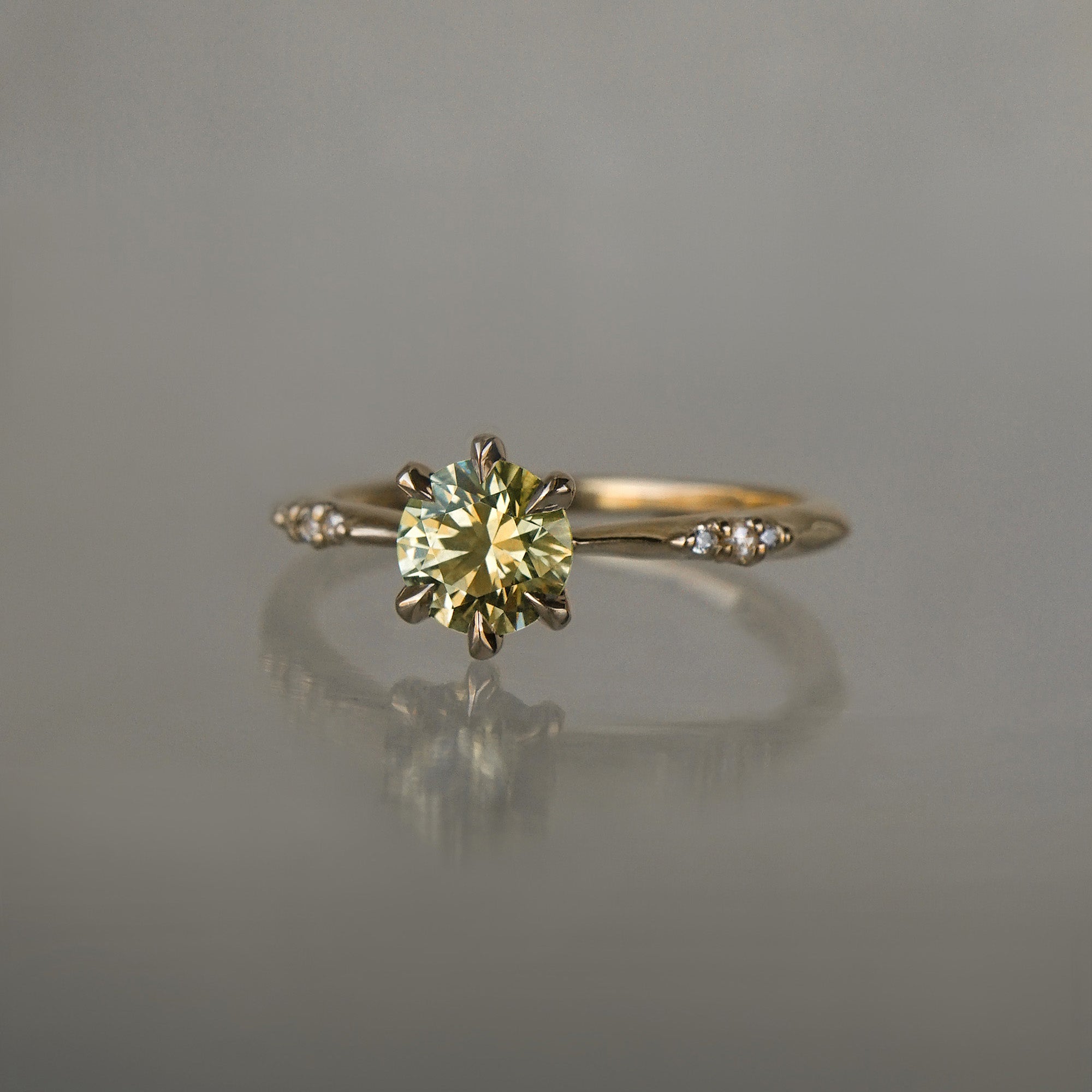 An elegant, one of a kind "Ilona" style engagement ring by Laurie Fleming Jewellery. The ring features a 1ct round brilliant cut yellow-grey Montana sapphire poised atop a tapered knife-edge band with small diamond accents on the band shoulders.  The ring is situated against a medium grey background.