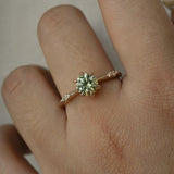 An elegant, one of a kind "Ilona" style engagement ring by Laurie Fleming Jewellery. The ring features a 1ct round brilliant cut yellow-grey Montana sapphire poised atop a tapered knife-edge band with small diamond accents on the band shoulders. The ring is worn on a hand against a plain light grey background.