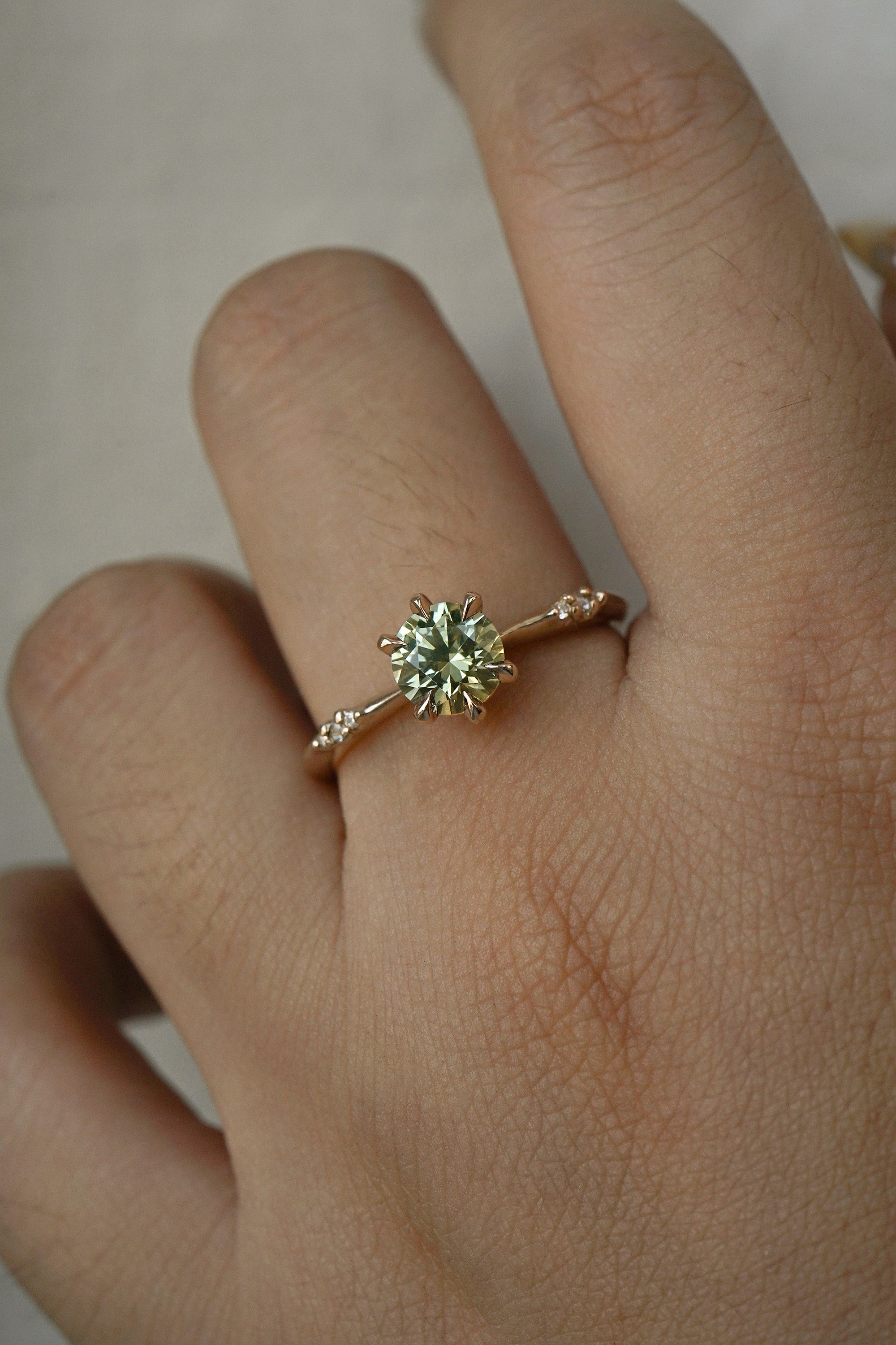 An elegant, one of a kind "Ilona" style engagement ring by Laurie Fleming Jewellery. The ring features a 1ct round brilliant cut yellow-grey Montana sapphire poised atop a tapered knife-edge band with small diamond accents on the band shoulders. The ring is worn on a hand against a plain light grey background.