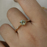 An elegant, one of a kind "Ilona" style engagement ring by Laurie Fleming Jewellery. The ring features a 1ct round brilliant cut yellow-grey Montana sapphire poised atop a tapered knife-edge band with small diamond accents on the band shoulders. The ring is worn on a hand against a plain light grey background.