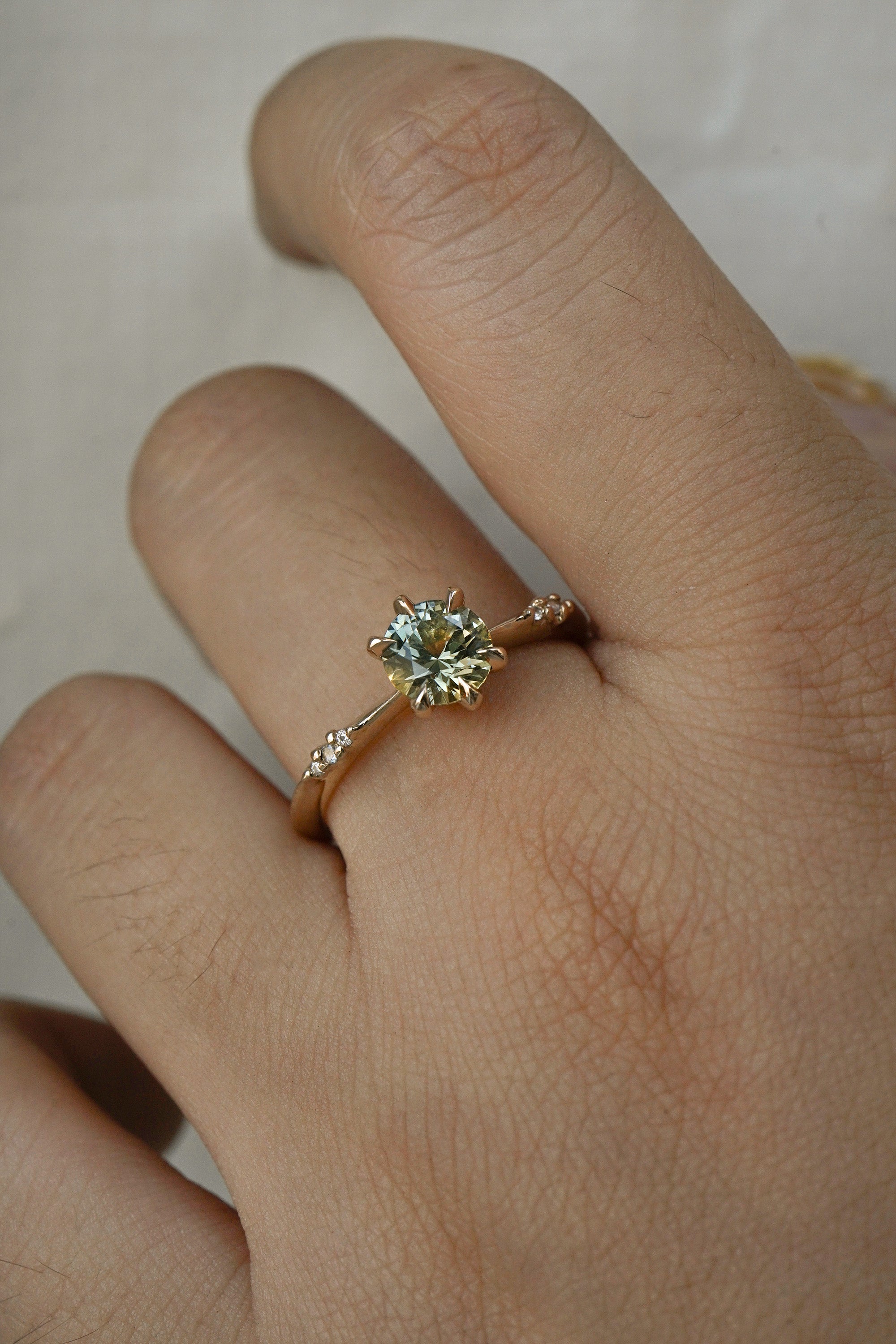An elegant, one of a kind "Ilona" style engagement ring by Laurie Fleming Jewellery. The ring features a 1ct round brilliant cut yellow-grey Montana sapphire poised atop a tapered knife-edge band with small diamond accents on the band shoulders. The ring is worn on a hand against a plain light grey background.